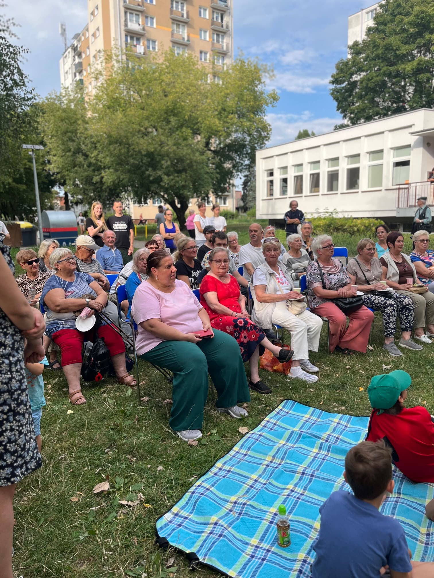 Mieszkańcy siedzą na kocach i krzesłach podczas prelekcji w plenerze