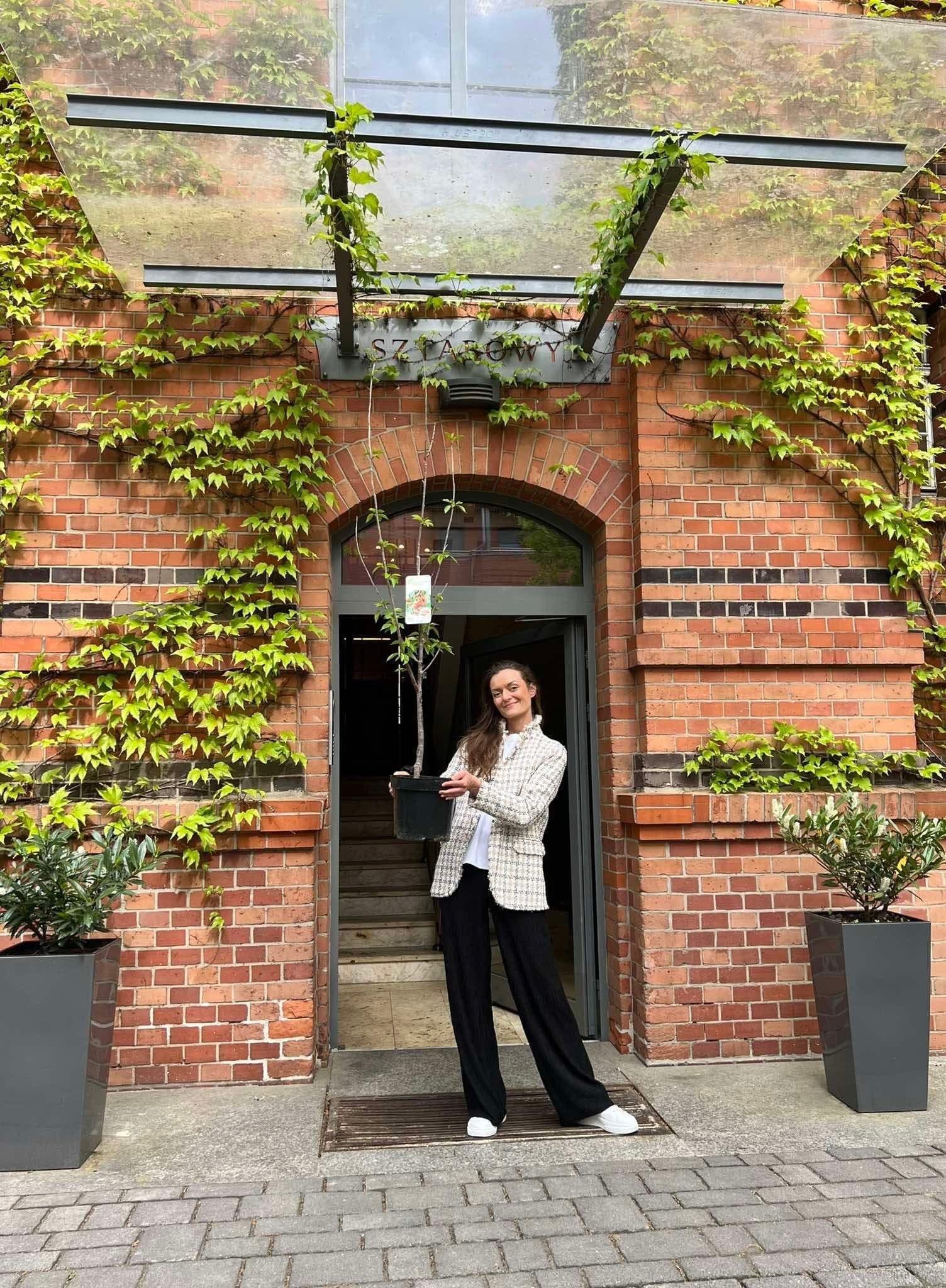 Kobieta trzymająca drzewko owocowe przed wejściem do budynku porośniętego bluszczem